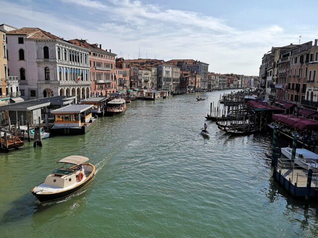 Blick von der Rialtobrücke auf den Canale Grande. Hier herrscht reger Verkehr sowohl auf den Wasserstraßen als auch auf den Uferpromenaden. Touristen füllen die sonnigen Gastgärten der Restaurants und Gondoliere werben  um Fahrgäste.