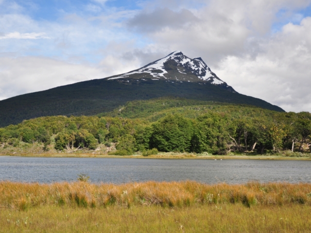 Den kleinen See umgibt eine Natur, die in prächtigen Farben schillert, während sich im Hintergrund sanft die schneebedeckten Berge erheben.